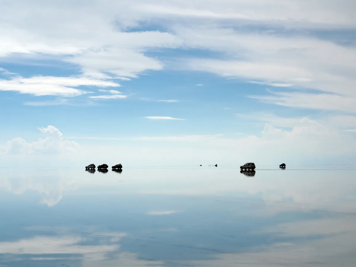 Private vehicles crossing a reflective salt flat beneath a wide blue sky